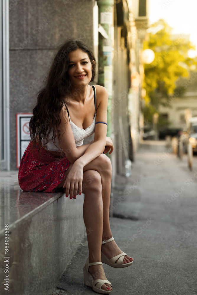 A young woman exudes a casual, relaxed charm as she sits on a ledge in an urban setting, wearing a summer dress and smiling gently. A young woman exudes a casual, relaxed charm as she sits on a ledge in an urban setting, wearing a summer dress and smiling gently.