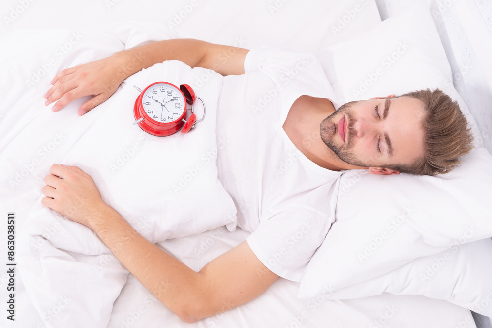 © be free - Morning wakeup. Sleepy man with alarm clock while lying in bed. Morning awakening. Sleepy man turning off alarm clock. Waking up early. Time to wakeup and get up. Lazy Sunday