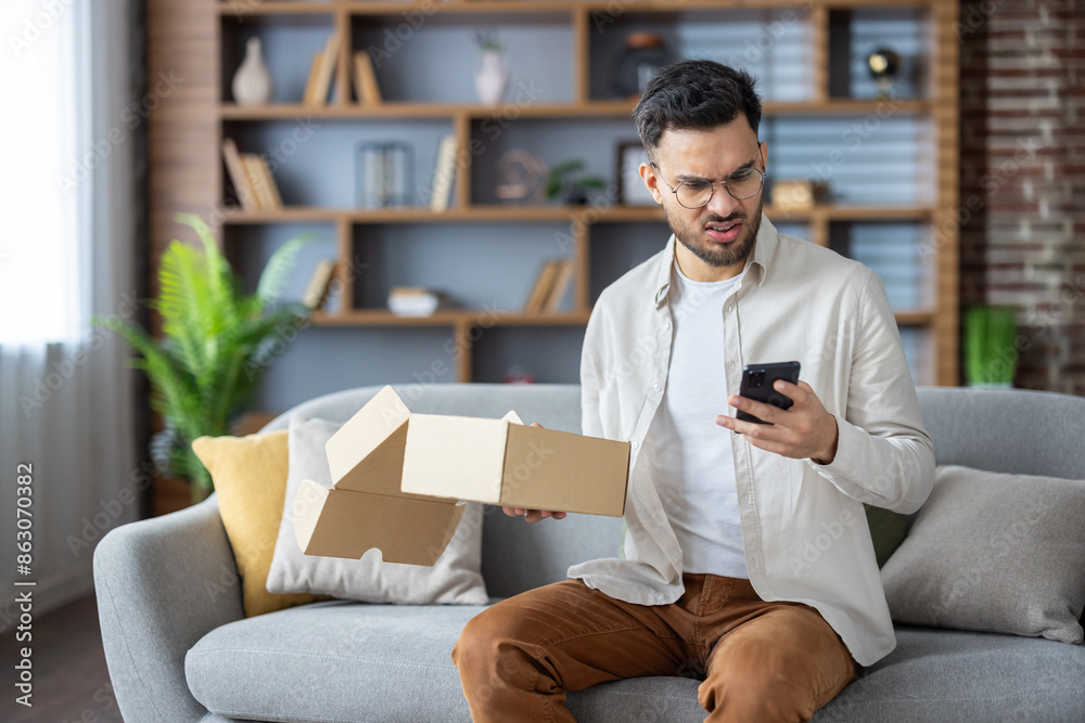 Obraz premium Angry young Indian man sitting on sofa at home, holding open parcel box and looking disappointedly at mobile phone, dialing delivery service with complaints
