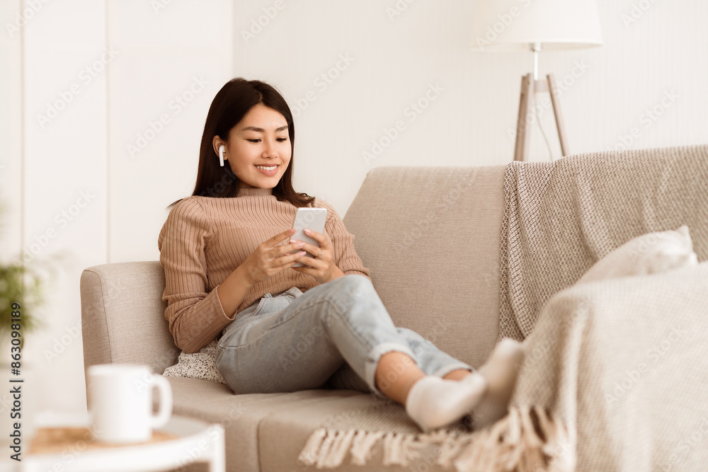 A woman is sitting comfortably on a couch with her feet up, using her smartphone. She is wearing a beige sweater and jeans. A woman is sitting comfortably on a couch with her feet up, using her smartphone. She is wearing a beige sweater and jeans.