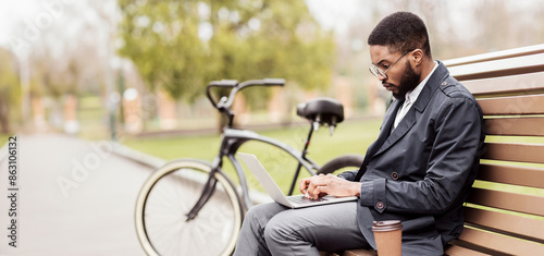 A business professional black man uses his laptop on a park bench beside his bike. The scene is calm and modern, highlighting a balance of work and relaxation in nature.
