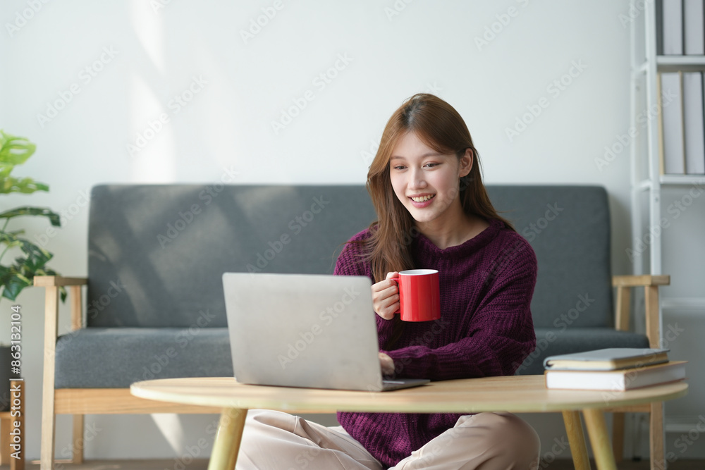 Smiling woman working on a laptop while holding a red coffee mug, seated at a wooden table in a cozy home setting. Smiling woman working on a laptop while holding a red coffee mug, seated at a wooden table in a cozy home setting.