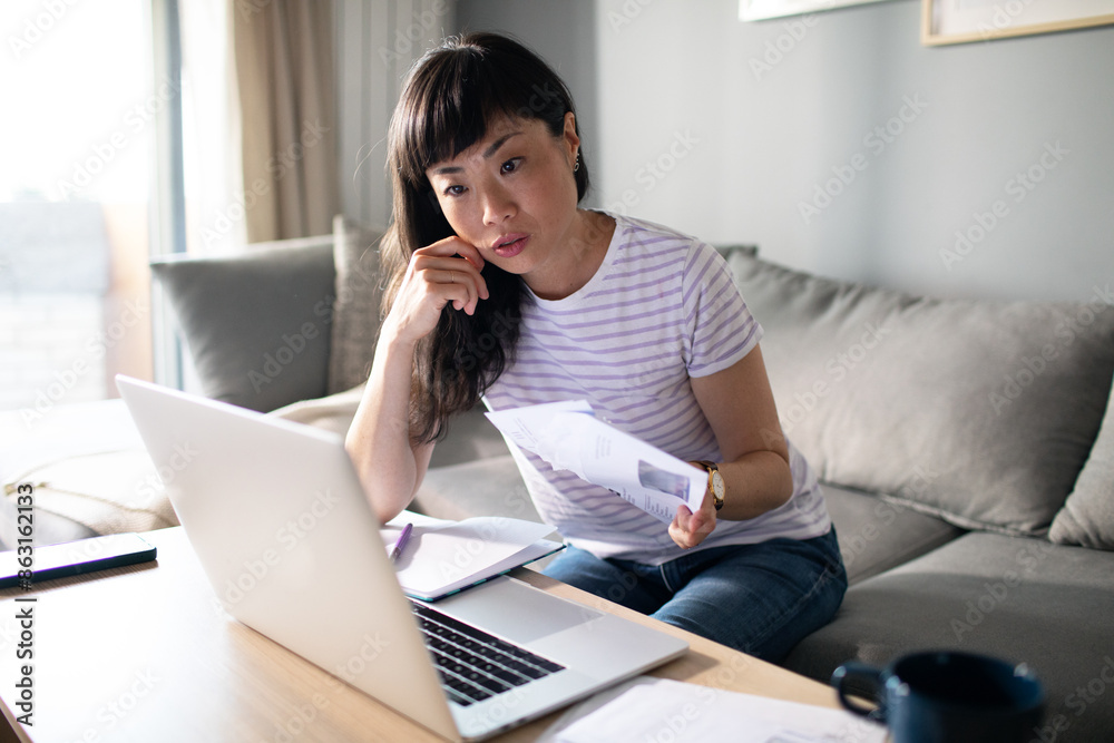 Concerned Asian woman holding bills and using laptop at home Concerned Asian woman holding bills and using laptop at home