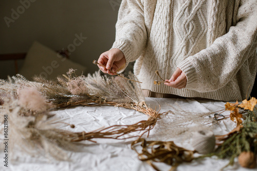 Woman making stylish autumn wreath. Hands with dried grass, herbs, leaves, ribbon and scissors on rustic table. Details of fall decor and arrangement in home
