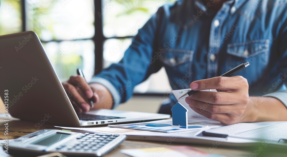 Real estate agent sits at his desk, looking over the realistic house model on top of papers with a calculator in front and holding an envelope in one hand while taking notes