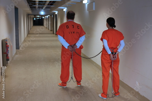 weibliche und männlicher Strafgefangene in Handschellen und Fusschellen und orangen Overall mit Sicherungskasten im Kellergang