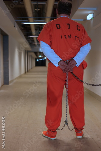 männlicher Strafgefangene in Handschellen und Fusschellen und orangen Overall mit Sicherungskasten im Kellergang