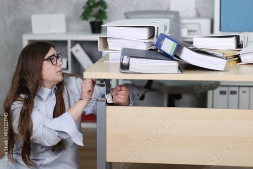 Sekretärin in Bluse, Hände in Handschellen, vor viele Aktenordnern im Büro, an den Schreibtisch gekettet, auf Boden sitzend