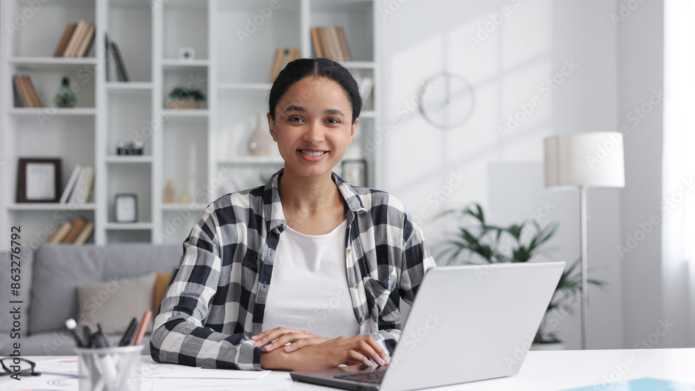 Portrait of a young African American woman working on a laptop at the table at home, turns her head, looks at the camera, smiles. Portrait of a young African American woman working on a laptop at the table at home, turns her head, looks at the camera, smiles.