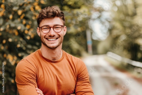 A happy man with glasses stands on a road, smiling with trees in the background. He is wearing an orange shirt and has his arms crossed.