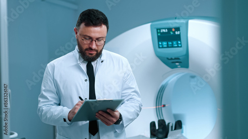 Experienced doctor standing at cabinet of magnetic resonance imaging. Tomographer holding tablet. Modern equipped room for MRI scanning. Doctor in gown at background of MRI capsule.