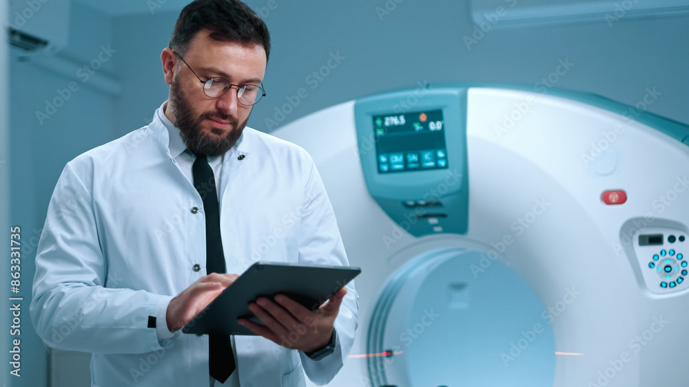 Obraz premium Experienced doctor standing at cabinet of magnetic resonance imaging. Tomographer holding tablet. Modern equipped room for MRI scanning. Doctor in gown at background of MRI capsule.