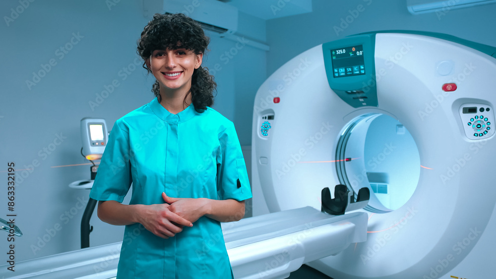 Young tomography specialist posing with wide smile. Cheerful doctor smiling at background of MRI equipment. Cabinet for MRI examination. MRI specialist. Tomography technician. Medical specialist. Young tomography specialist posing with wide smile. Cheerful doctor smiling at background of MRI equipment. Cabinet for MRI examination. MRI specialist. Tomography technician. Medical specialist.