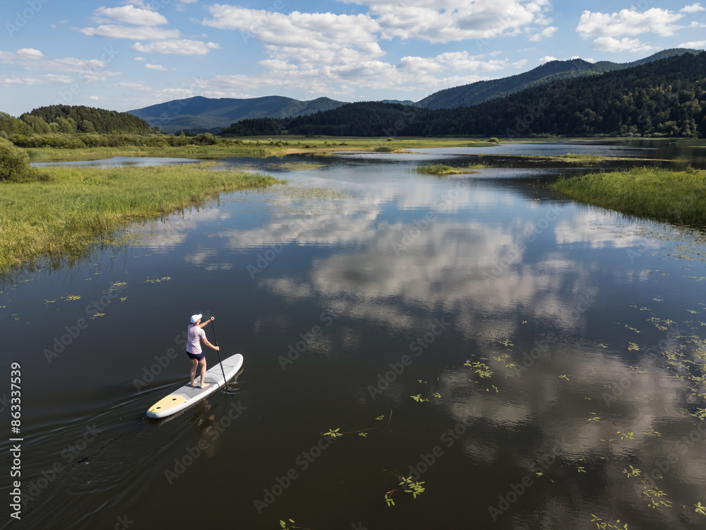 Woman paddling stand up board, relaxing summer activity in lake natural surroundings, aerial shot. Relaxation, fitness, and exploration concepts. Woman paddling stand up board, relaxing summer activity in lake natural surroundings, aerial shot. Relaxation, fitness, and exploration concepts.