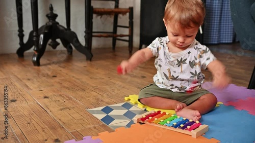Cute baby boy knocking on colorful iron plates, learning playing educational music toy, Toddler plays with colorful xylophone and educational toys while sitting on a mat indoors. Learning through play