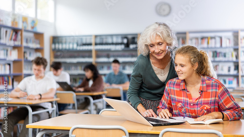 Mature female teacher working with schoolchildren in the library cheks the assignment from a European fifteen-old-year schoolgirl photo