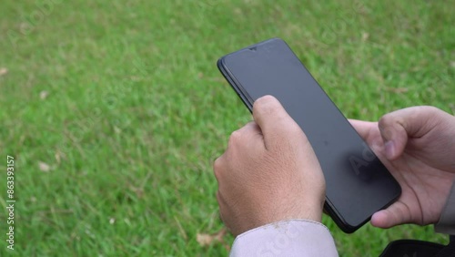 Someone is sitting in the middle of green and beautiful nature, surrounded by fresh grass. The person looks engrossed in playing on his cellphone