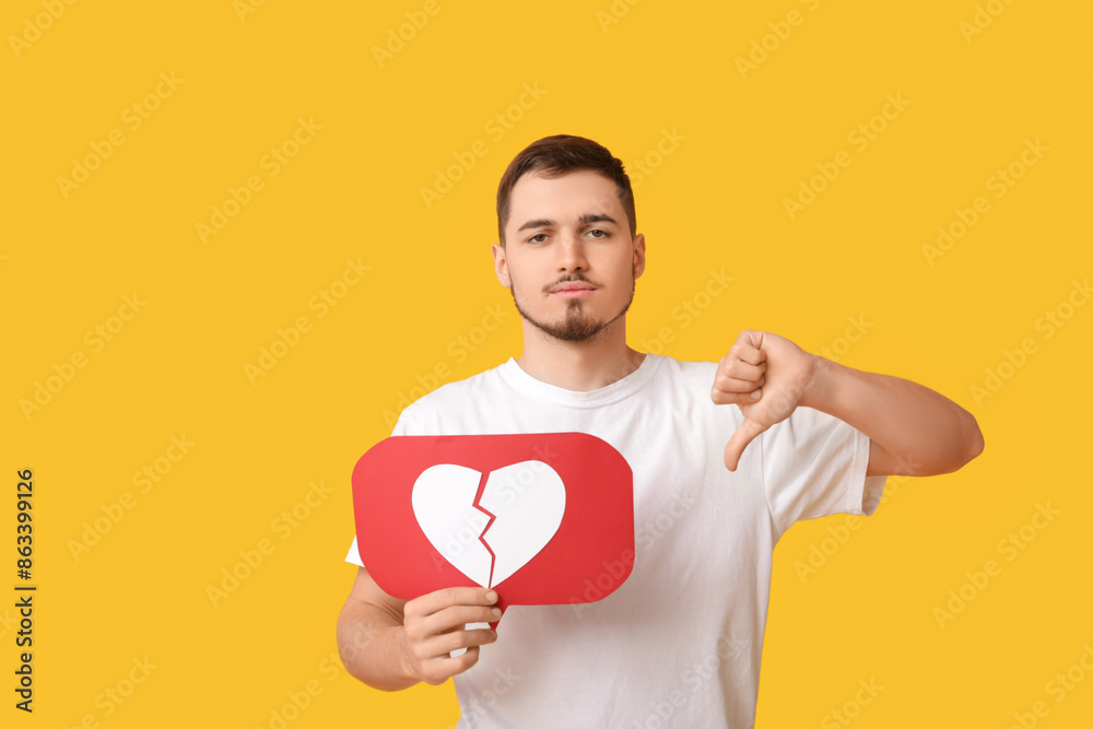 Handsome young displeased man with heart dislike icon showing thumb-down gesture on yellow background Handsome young displeased man with heart dislike icon showing thumb-down gesture on yellow background