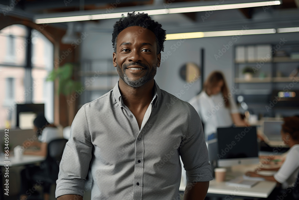 Confident African American businessman in casual attire smiling in modern tech office with team working behind Confident African American businessman in casual attire smiling in modern tech office with team working behind