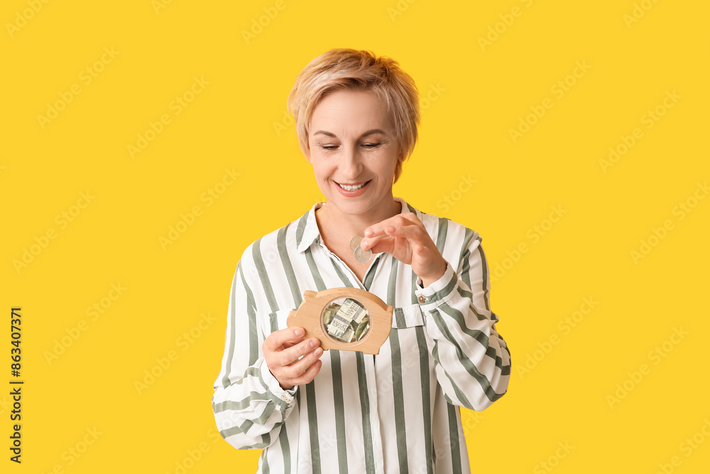 Beautiful mature woman putting coins into wooden piggy bank on yellow background Beautiful mature woman putting coins into wooden piggy bank on yellow background