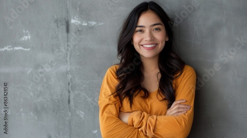 A young Hispanic woman smiles pleasantly while standing with crossed arms against a grey wall. She is looking directly at the camera. Generative AI