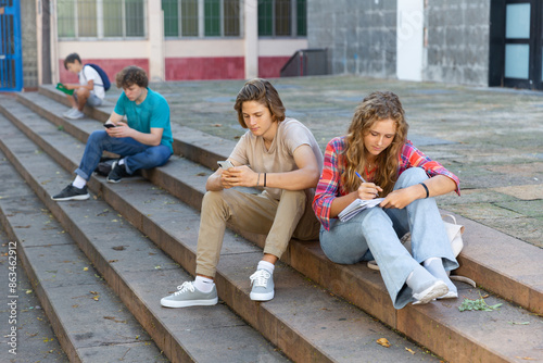 Teenagers sitting on stairs in school campus and doing their own things. Girl preparing for exams and her classmate sitting next to her and using smartphone.
