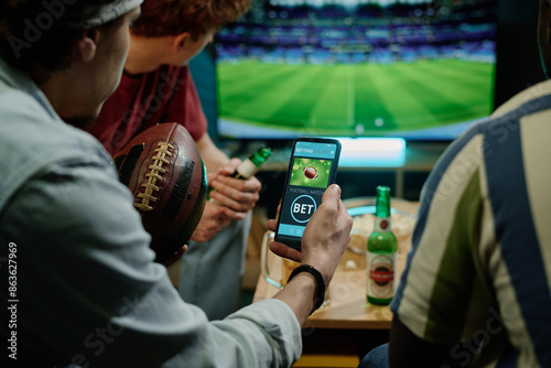 Crop shot of young male friends betting on team in app on smartphone while watching football match on TV