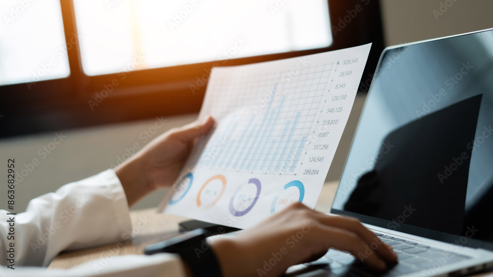 A person is holding a piece of paper with graphs on it and typing on a laptop. Concept of analyzing data and making informed decisions based on the information presented. Scene is focused and serious A person is holding a piece of paper with graphs on it and typing on a laptop. Concept of analyzing data and making informed decisions based on the information presented. Scene is focused and serious