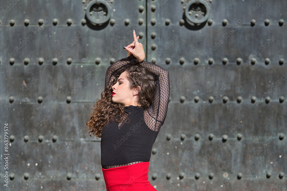 Fototapeta premium Portrait of young, beautiful, brunette woman in black shirt and red skirt, dancing flamenco with old, black metal door in the background. Flamenco concept, dance, art, typical Spanish.