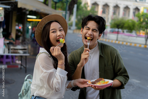 Asian couple traveler wearing a casual wear and hat in relationship buying a dessert snack on street food market in the evening. Backpacker traveling in Bangkok, Thailand..