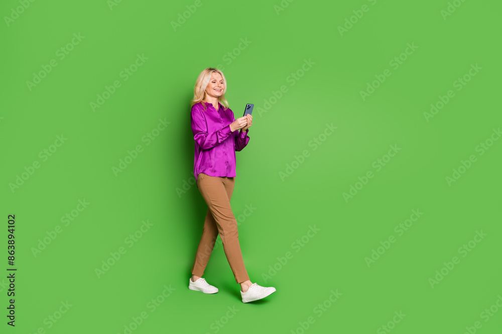 Full size photo of attractive retired woman hold device walking dressed stylish violet satin clothes isolated on green color background