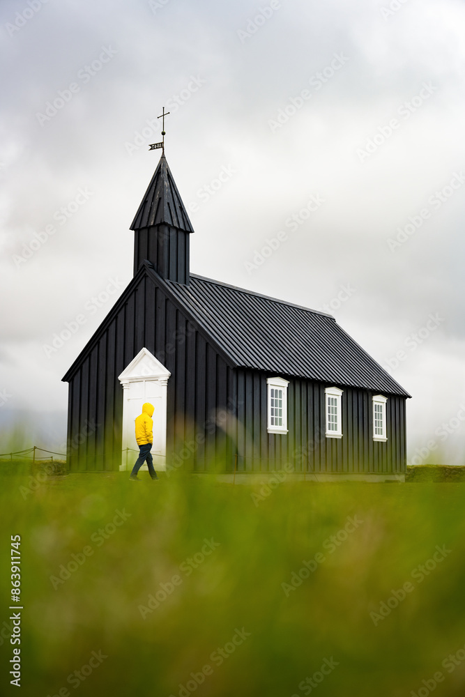 Stunning view of a tourist wearing a yellow jacket passing in front of the black church of Budir. Búðakirkja is a small wooden church located on the south side of Snæfellsnes, Iceland.