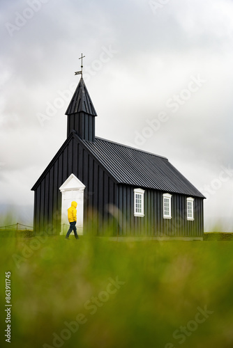 Obraz na płótnie Stunning view of a tourist wearing a yellow jacket passing in front of the black church of Budir. Búðakirkja is a small wooden church located on the south side of Snæfellsnes, Iceland. w salonie