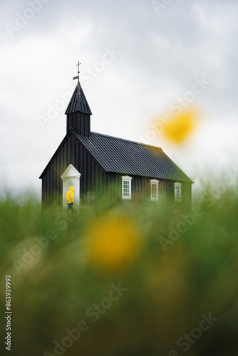 Obraz na płótnie Stunning view of a tourist wearing a yellow jacket passing in front of the black church of Budir. Búðakirkja is a small wooden church located on the south side of Snæfellsnes, Iceland. w salonie