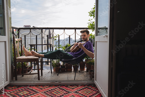 A man plays the ukulele on a balcony, surrounded by nature in the calm morning ambiance