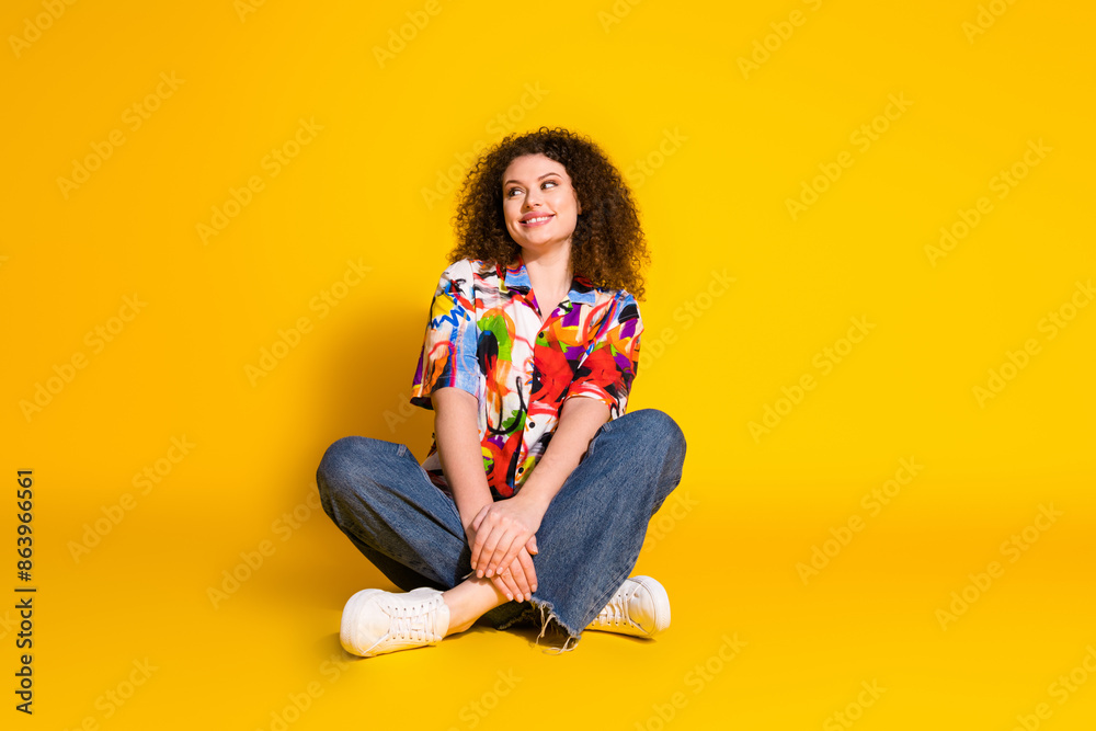 Full body photo of pretty young girl sit floor look empty space wear trendy colorful print outfit isolated on yellow color background Full body photo of pretty young girl sit floor look empty space wear trendy colorful print outfit isolated on yellow color background