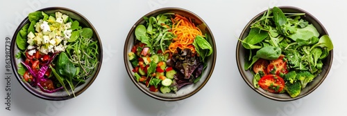 Overhead view of three colorful salad bowls with fresh vegetables and greens, arranged on a white surface