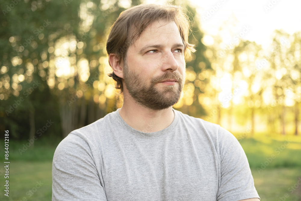 Portrait of handsome young man in gray t-shirt outdoors. Male with long hair and beard feel happy, calm and serious on Green nature sunset background. Man looking away into distance. Diversity People Portrait of handsome young man in gray t-shirt outdoors. Male with long hair and beard feel happy, calm and serious on Green nature sunset background. Man looking away into distance. Diversity People
