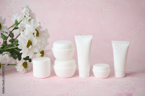 White tubes and jars of cream with bouquet of flowers on a pink background