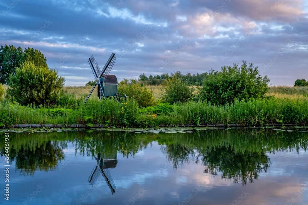 Obraz premium Dutch windmill, green fields, beautiful evening sky, clouds reflecting in the water.