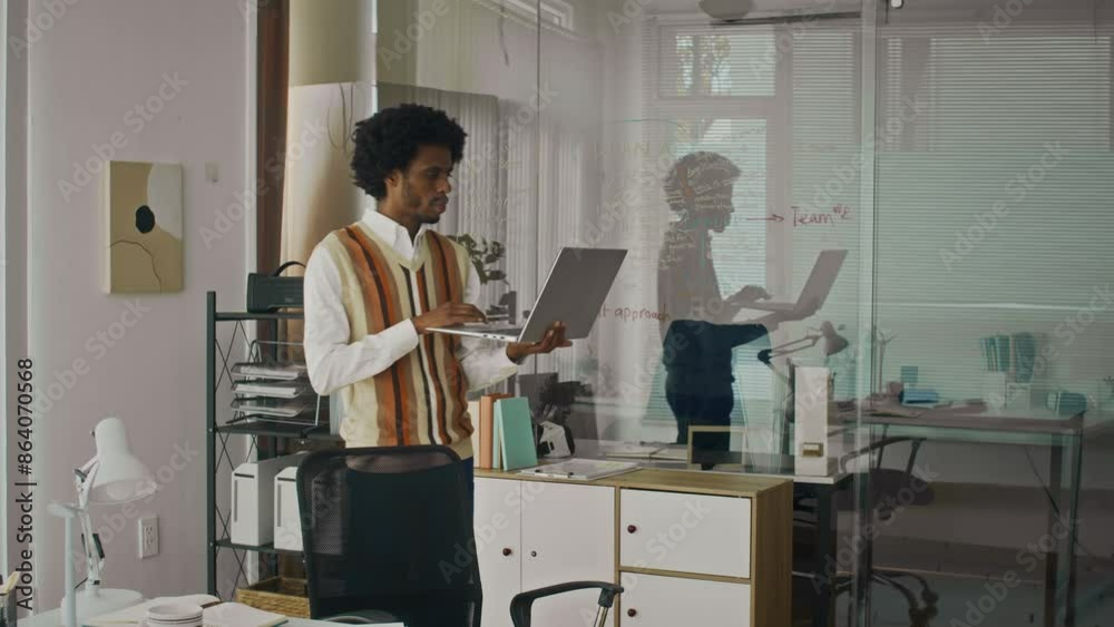 Medium long shot of African American product manager holding wireless laptop while thinking about ideas of project at startup office Medium long shot of African American product manager holding wireless laptop while thinking about ideas of project at startup office
