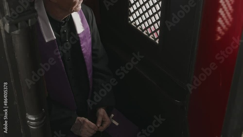 Tilt up shot of senior priest in black robe and purple stole sitting inside of confession booth and looking through lattice screen
