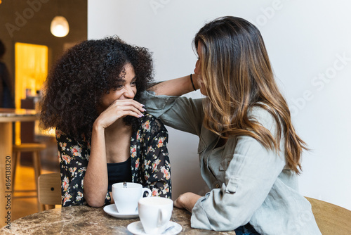 Smiling diverse girlfriends interacting at table in coffee house. Cheerful female putting elbow on shoulder of ethnic friend while talking at cafeteria table with cups of coffee