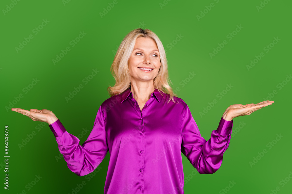 Photo portrait of nice senior female look compare hold empty space wear trendy violet satin outfit isolated on green color background Photo portrait of nice senior female look compare hold empty space wear trendy violet satin outfit isolated on green color background