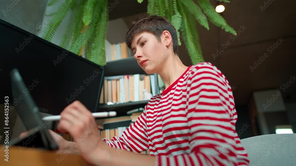 Camera moving around Caucasian girl with short dark hair. Woman creating illustrations with special pen on tablet device. Big monitors in front of female. Working remotely from home.