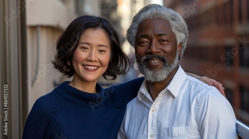 Middle-aged Asian woman in blue sweater and happy black man in white shirt standing on a balcony in afternoon light