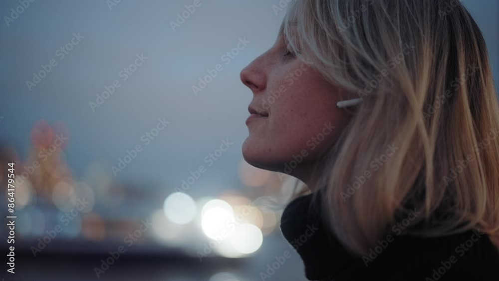 Blonde woman with headphones by the river in New York. Young woman enjoying evening walk by the Hudson river with light reflections in the background. Blonde woman with headphones by the river in New York. Young woman enjoying evening walk by the Hudson river with light reflections in the background.