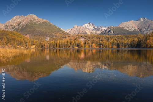 Autumn view of the Tatras in Slovakia: golden, red, and orange leaves on trees surround majestic mountains with snow-capped peaks. Clear, blue sky contrasts with the warm colors of the forest. 