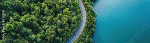 Aerial view of a winding road through dense green forest next to a calm blue lake, showcasing nature's harmony and beauty.