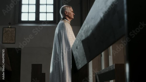 Side view of senior pastor dressed in white chasuble walking among empty pews in Lutheran church